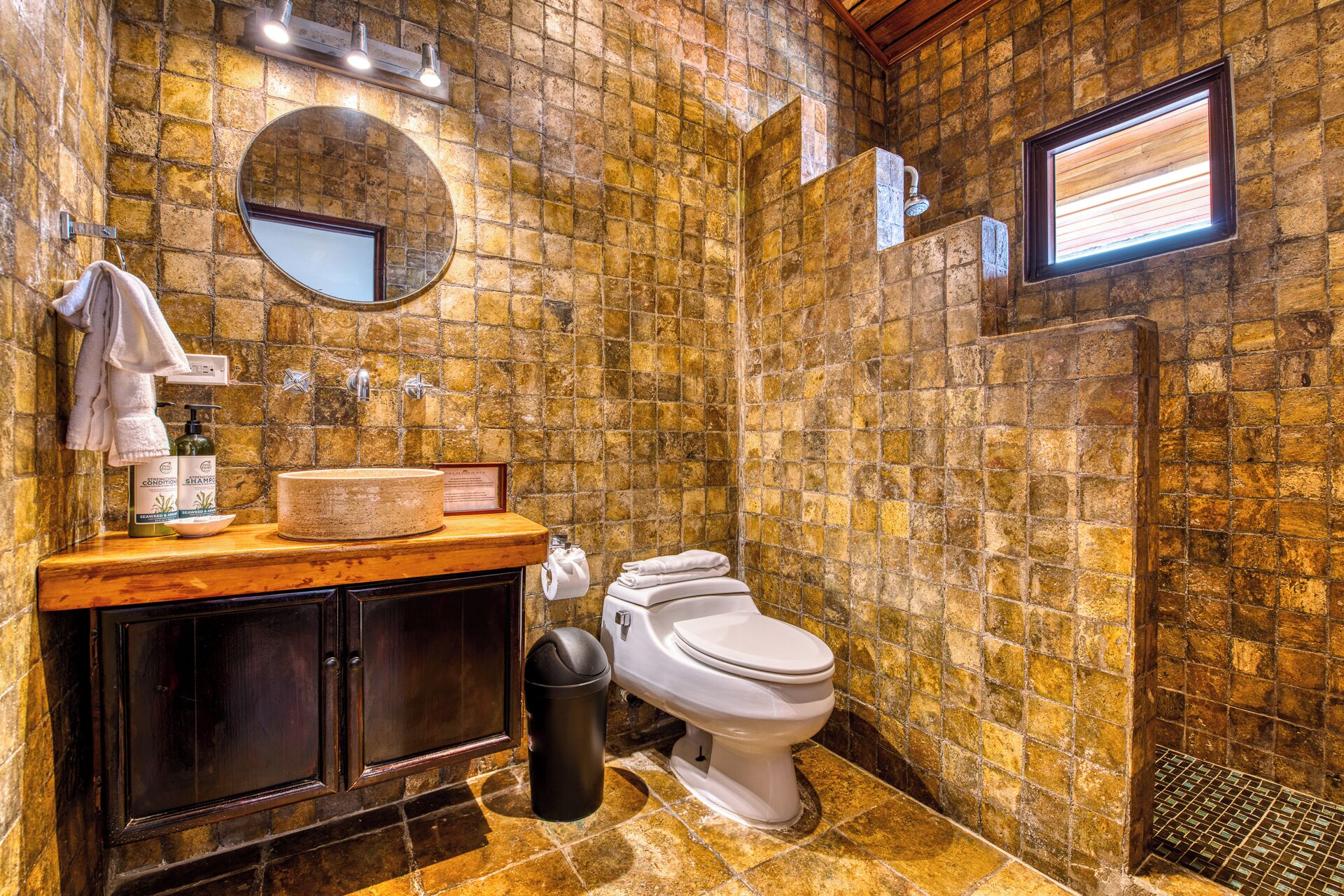 Un baño con pared de piedra, un espejo redondo, un tocador de madera, un inodoro y una ventana.