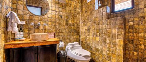 Un baño con pared de piedra, un espejo redondo, un tocador de madera, un inodoro y una ventana.