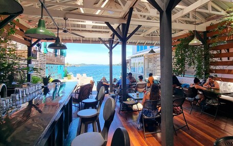 A bar area with outdoor seating overlooking a body of water, featuring wooden beams and hanging lights.