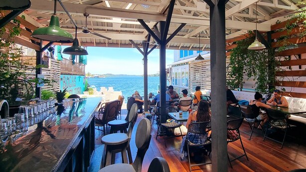 A bar area with outdoor seating overlooking a body of water, featuring wooden beams and hanging lights.