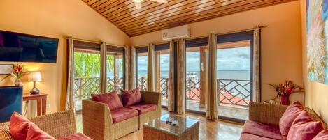 A spacious living area with wicker furniture, a ceiling fan, and large windows offering a view of the sea.