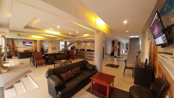 Um amplo hall de hotel com balcão de recepção, área de estar e escada que dá acesso aos andares superiores.