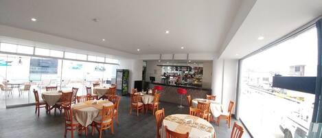 A spacious indoor dining area with multiple tables and chairs, a bar area in the background, and large windows providing a view of the outside.
