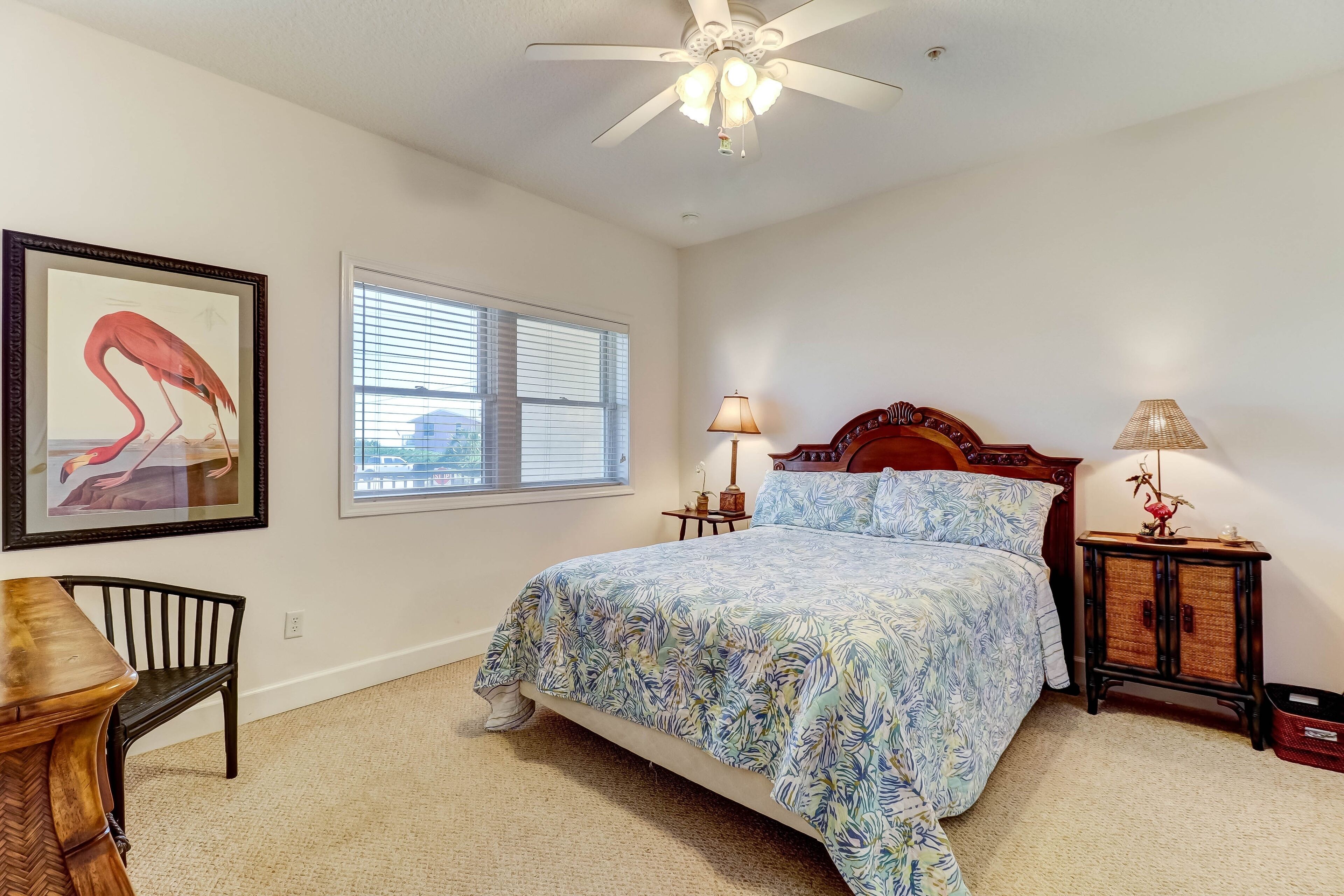 A bedroom with a bed, a ceiling fan, a window with blinds, a framed picture, a nightstand with a lamp, and a chair.