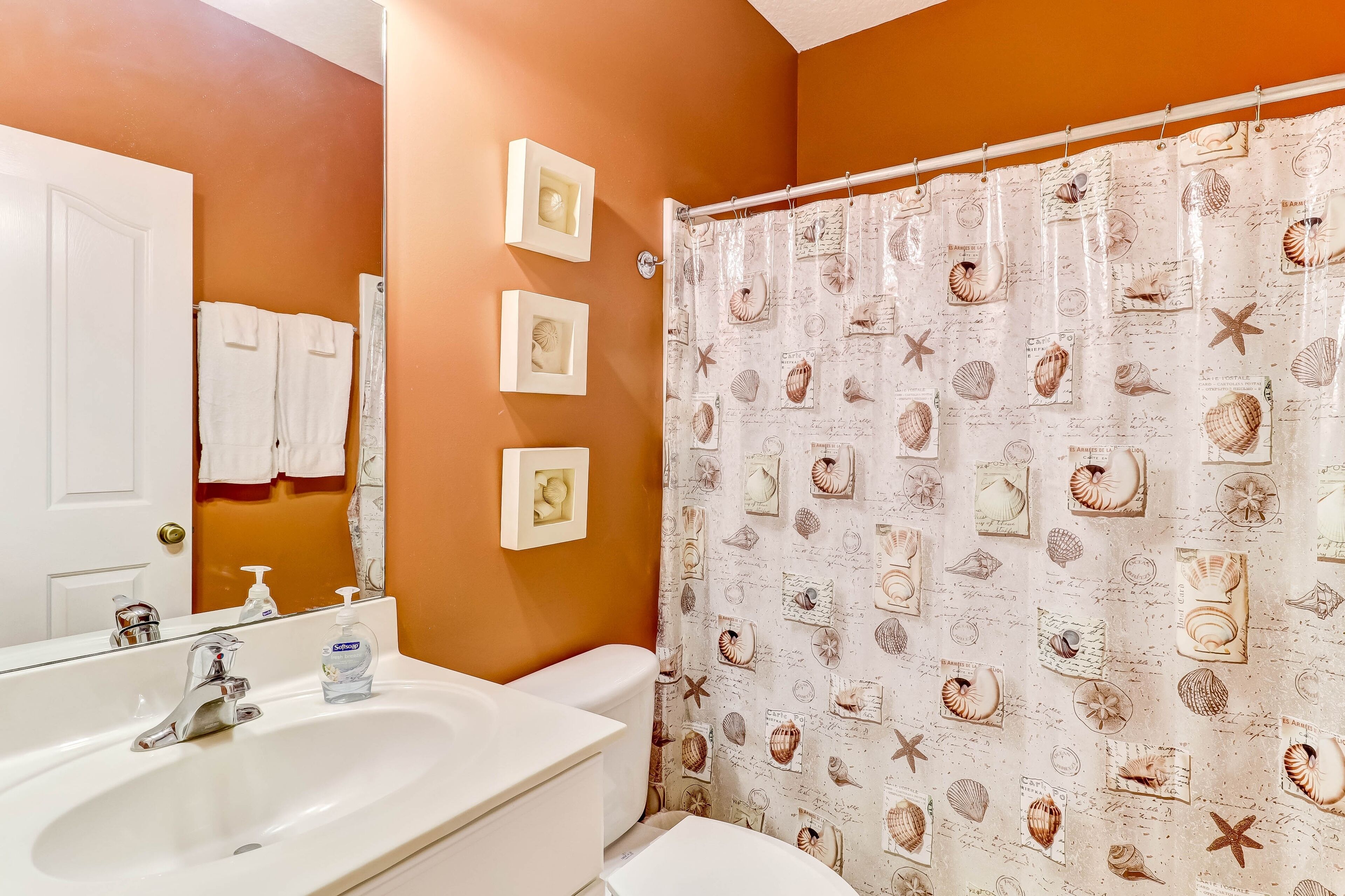 A bathroom with a white sink, a toilet, and a shower curtain with a sea shell pattern. There are three framed pictures on the wall.