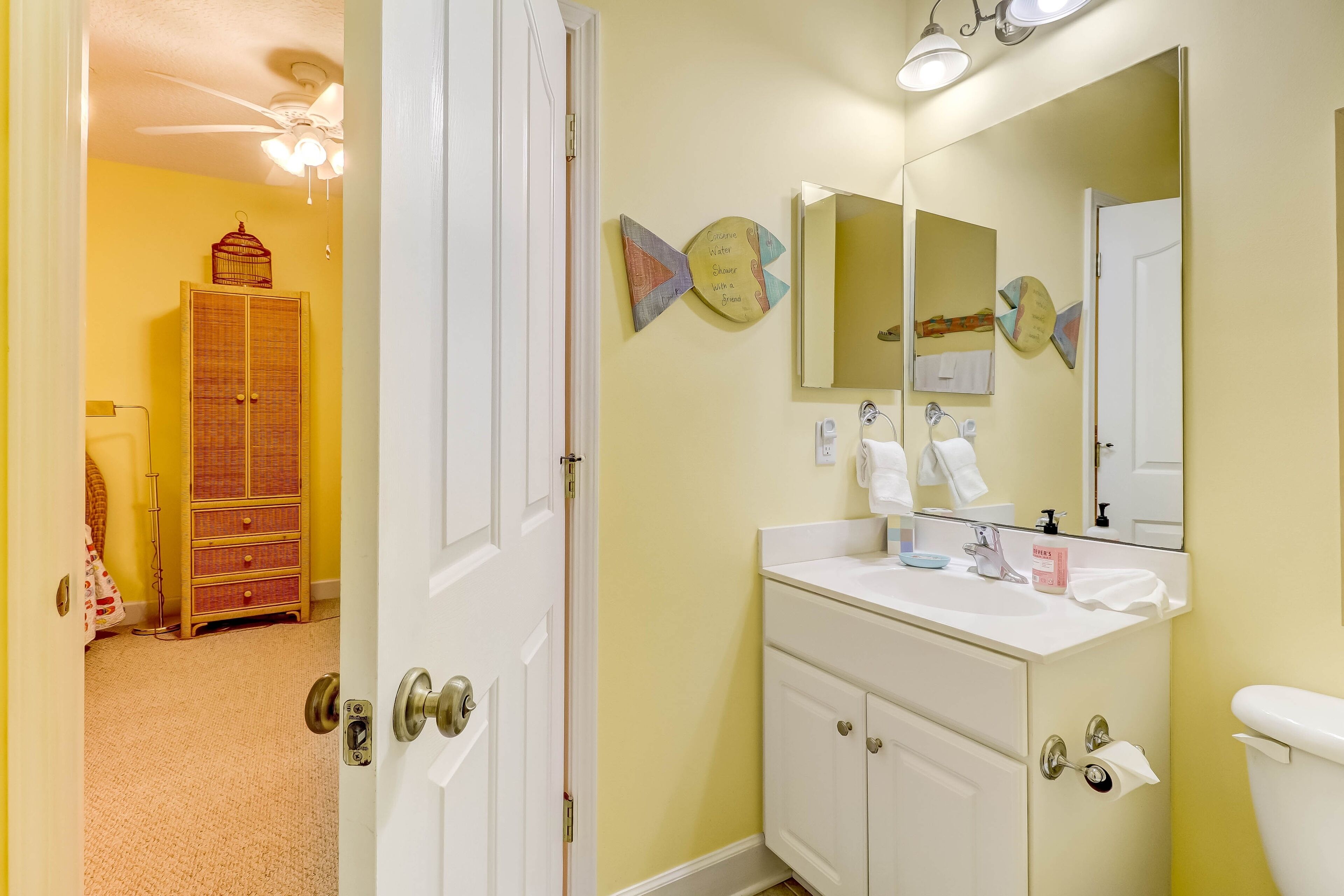 A bathroom with a white vanity, a large mirror, and a toilet. There is a wooden cabinet and a ceiling fan in the adjacent room.