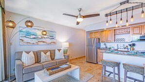 A modern living room with a grey sofa, a coffee table, and a kitchen area with wooden cabinets and a refrigerator.