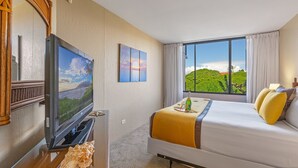 A hotel room with a bed, a television, a window with a view of greenery, and a painting on the wall.