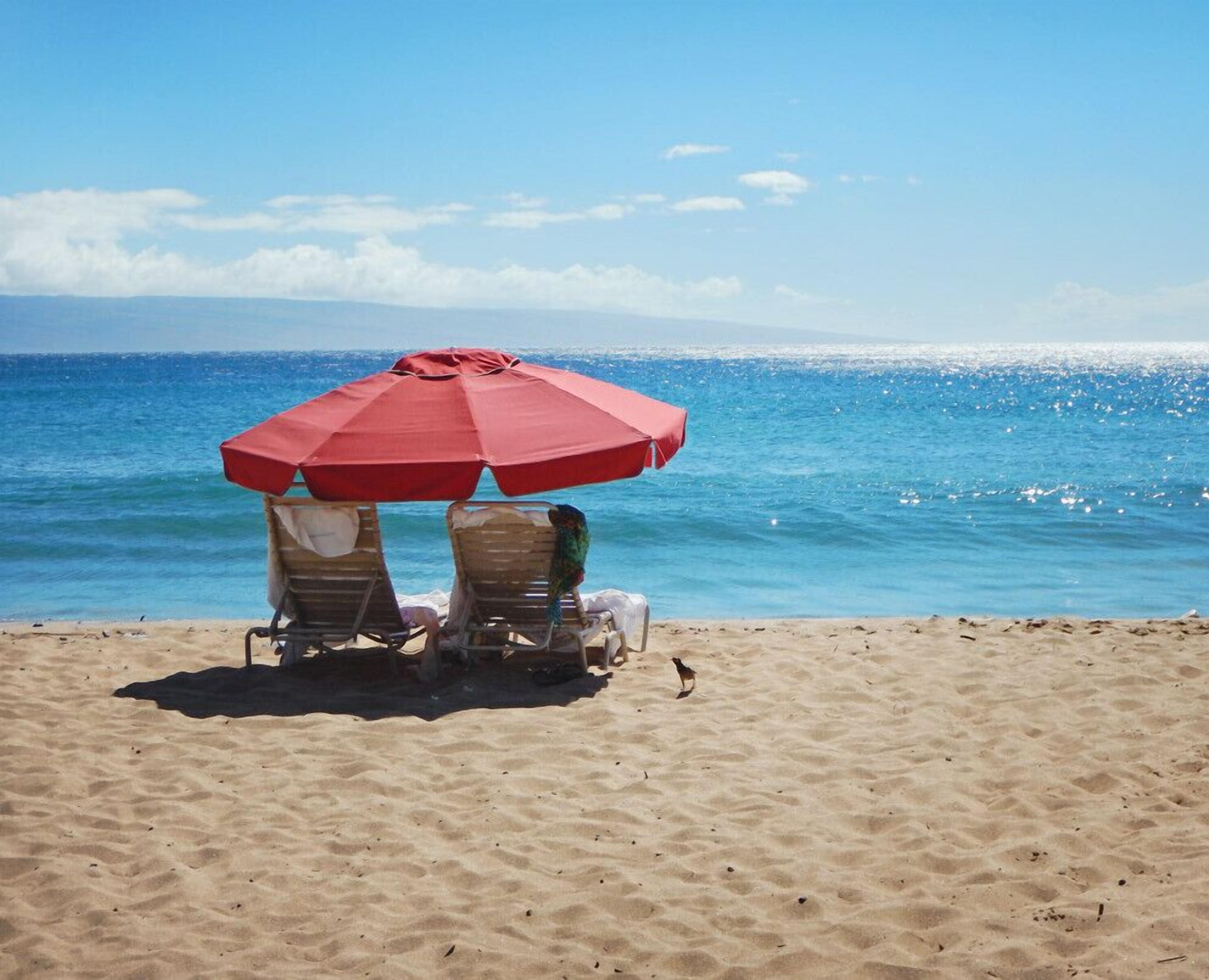 Deux chaises de plage sous un parasol rouge, sur une plage de sable, avec l’océan en arrière-plan.
