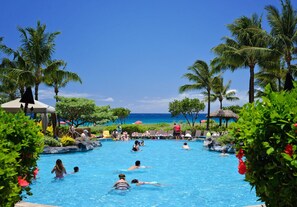 Une piscine entourée de palmiers, avec des personnes qui nagent et se détendent, et d’où l’on peut admirer la vue sur l’océan.