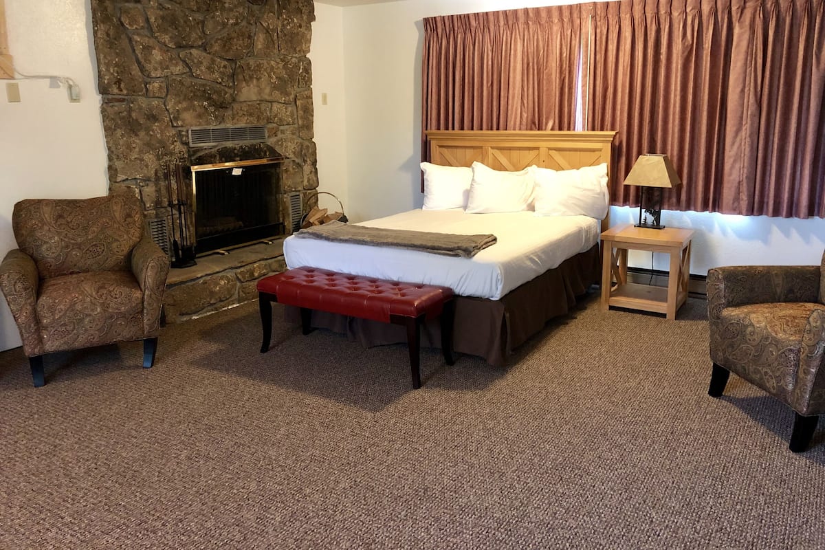 A bedroom with a stone fireplace, a bed with white linens, a red bench, and two patterned armchairs.