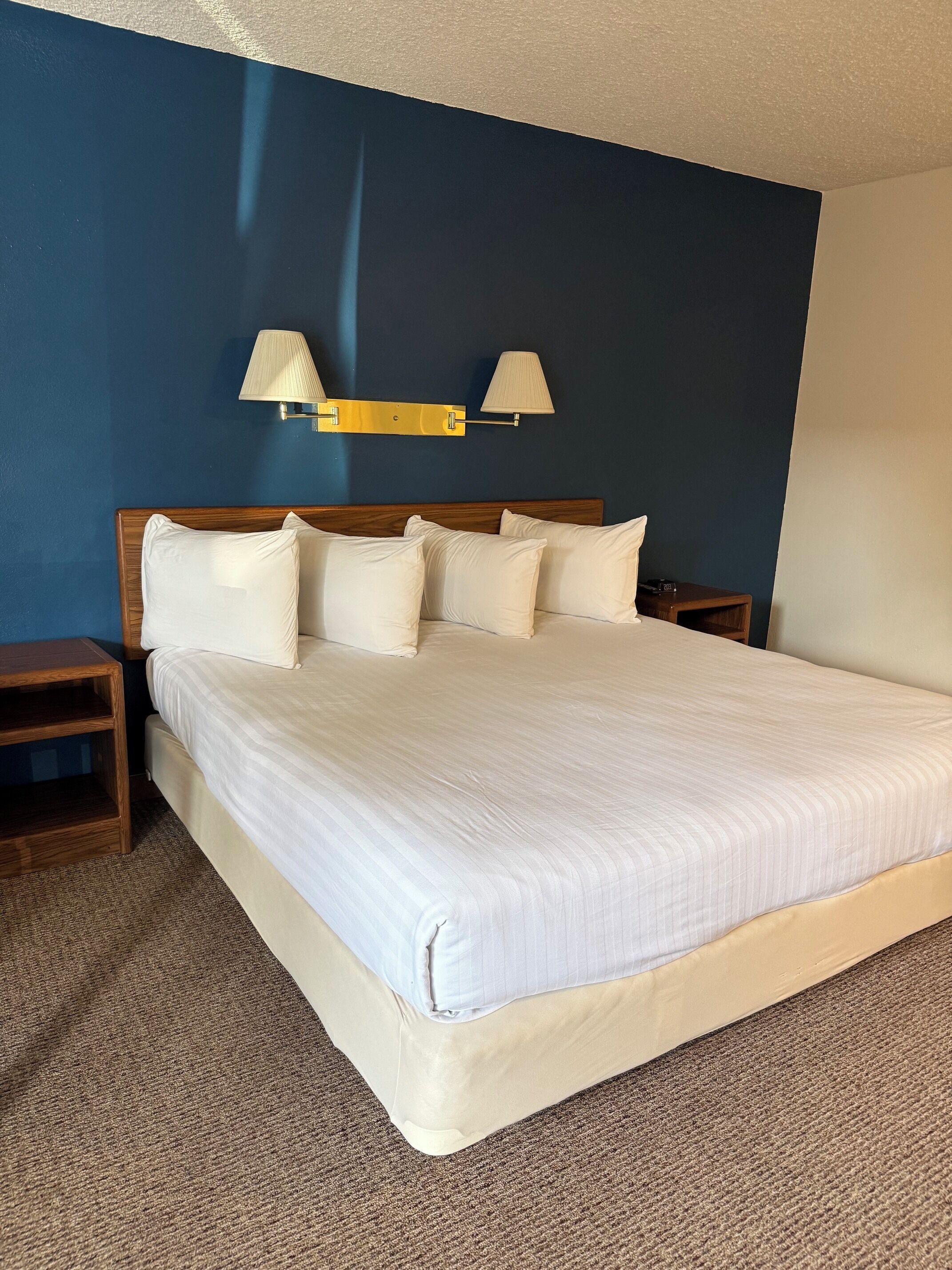 A hotel room with a large bed, two bedside lamps, a wooden nightstand, and a carpeted floor.