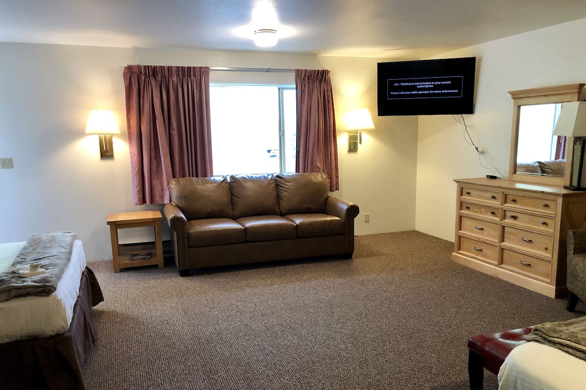 A hotel room with a brown leather sofa, a wooden dresser, a bed with a white comforter, and a flat-screen TV mounted on the wall.