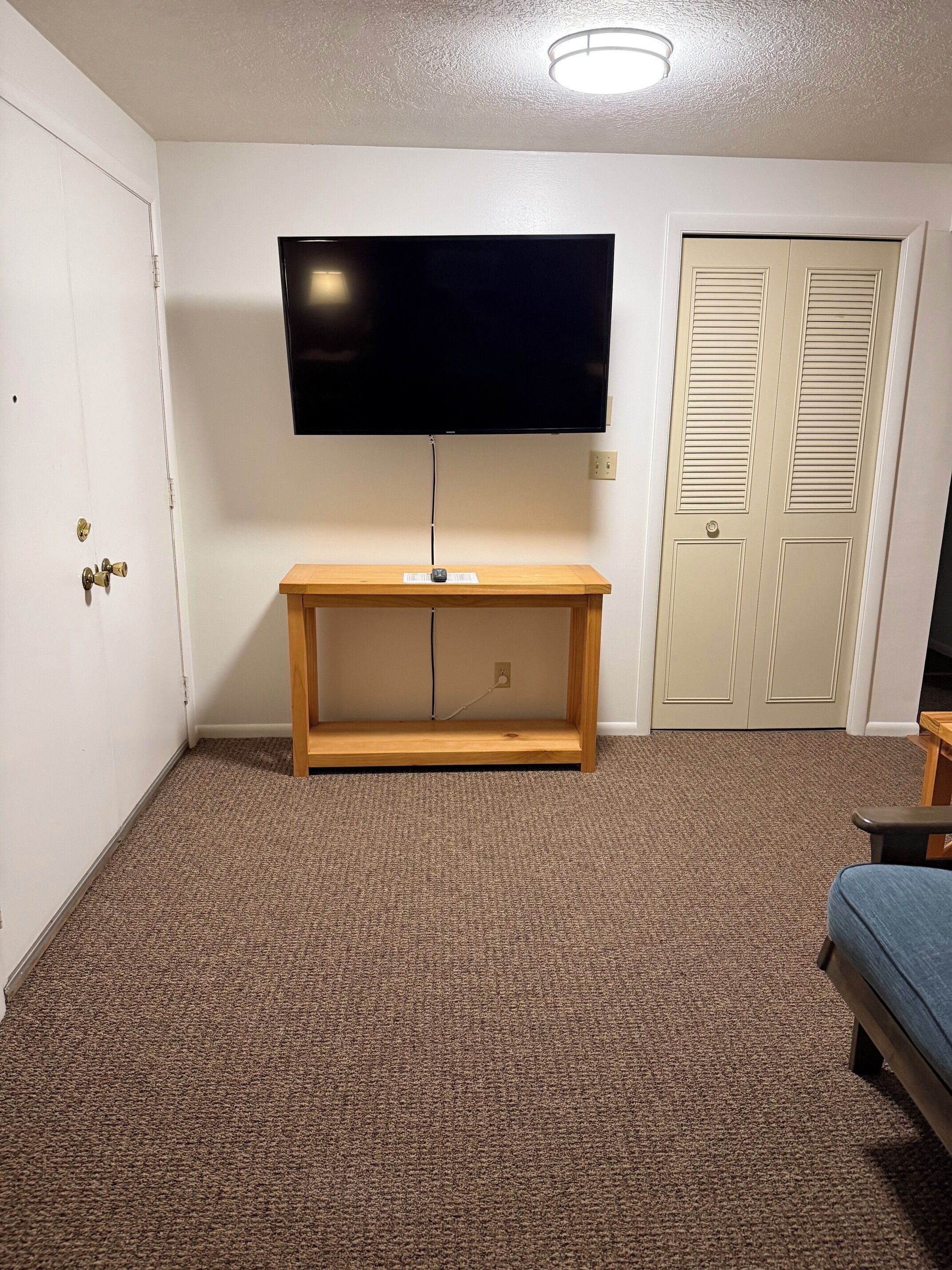A room with a television mounted on the wall, a wooden bench, a carpeted floor, a door with a lock, and a closet with louvered doors.