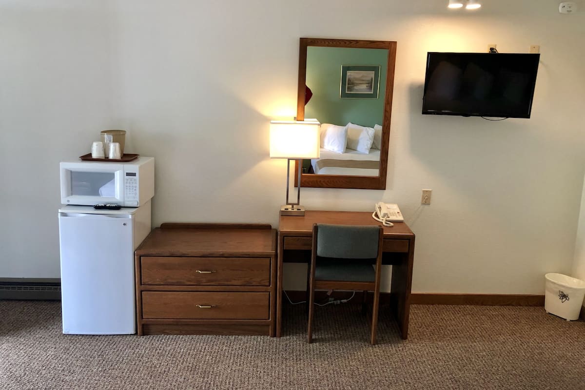 A hotel room with a desk, a microwave, a refrigerator, a lamp, a telephone, a TV, and a trash can.
