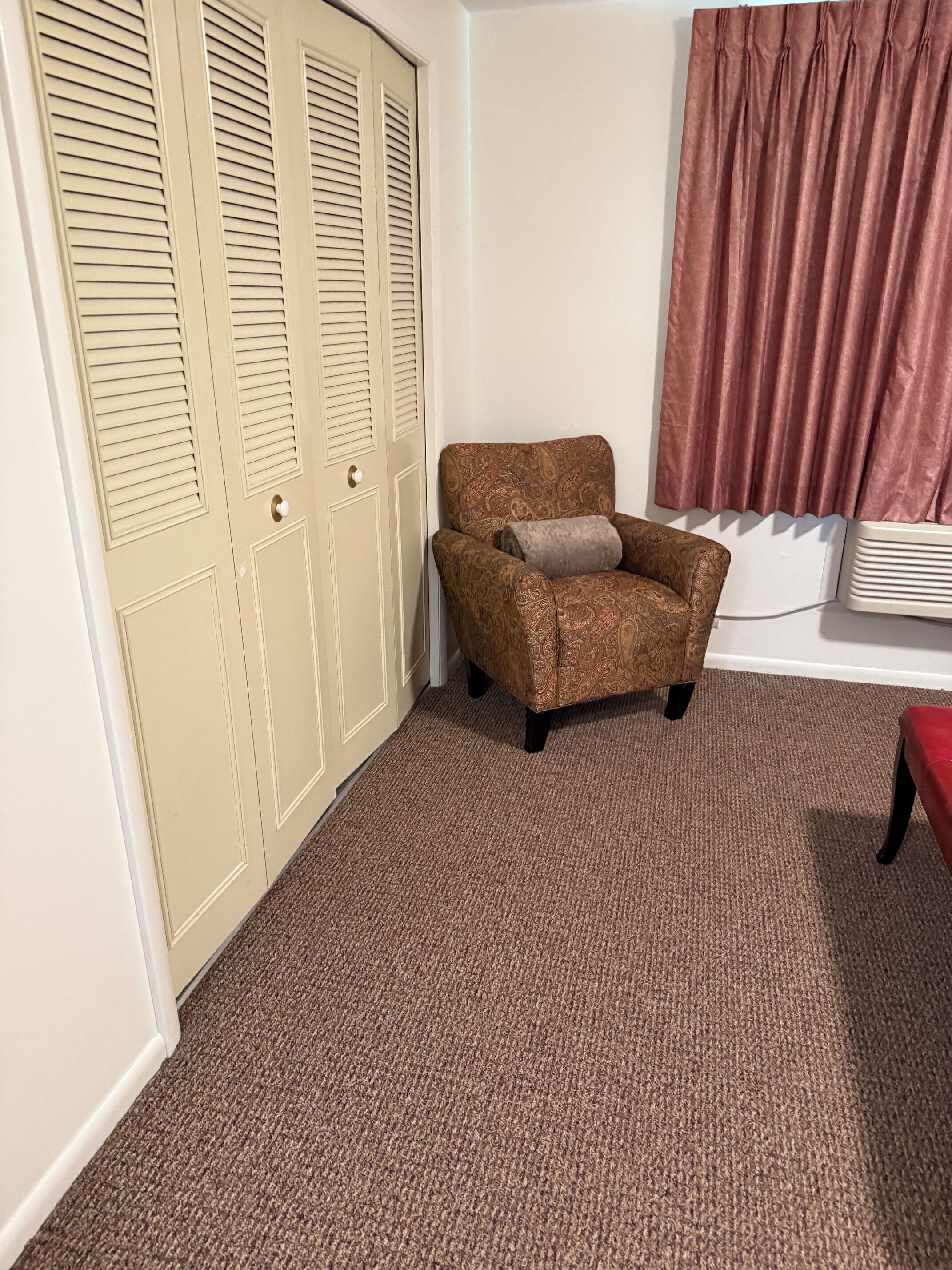 A room with a brown patterned armchair, beige louvered closet doors, a brown carpet, and a red curtain.