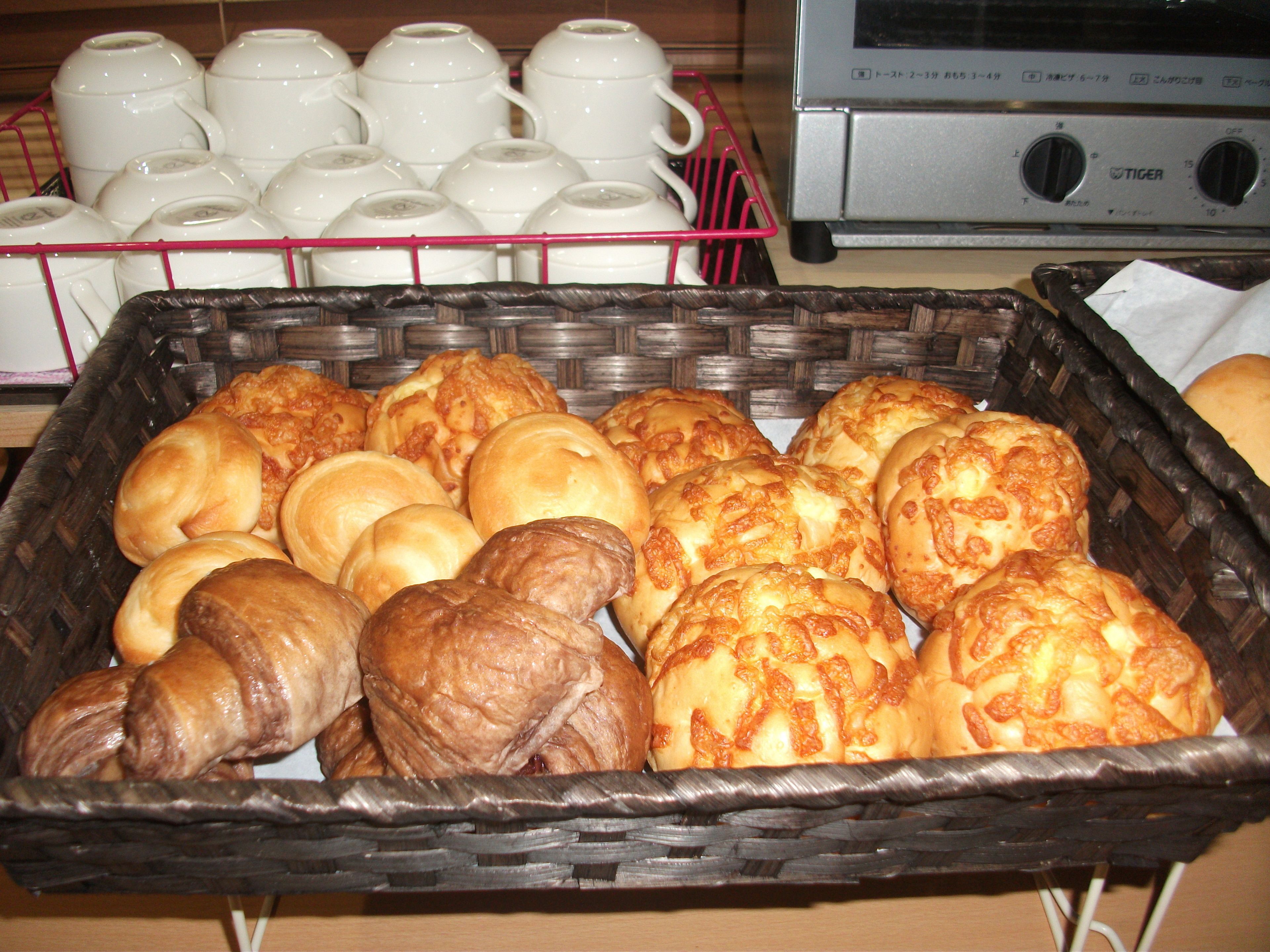A basket of assorted baked goods, a rack of white ceramic cups, and a silver appliance with Japanese text.