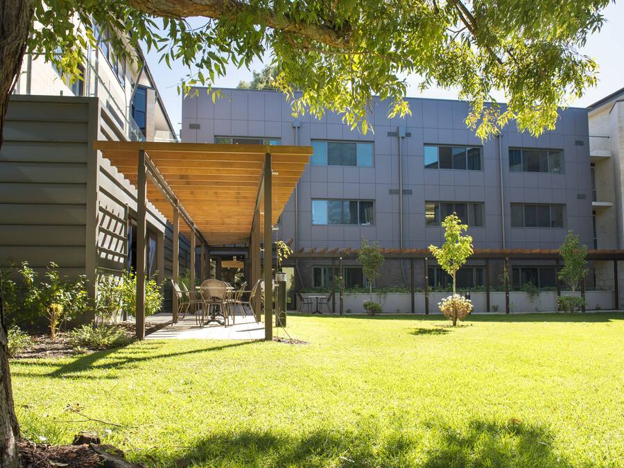 Un patio con un pergolado de madera, mobiliario exterior y un edificio con múltiples ventanas y balcones.