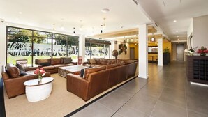 A spacious lobby with a large brown leather sofa, a white coffee table, and a large window offering a view of parked cars outside. - St Catherine's on Park (Crawley)