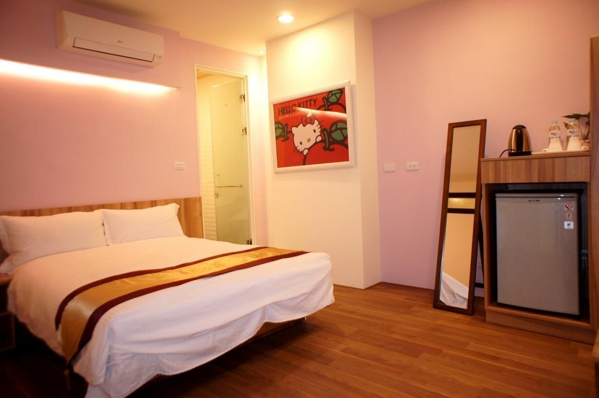 A hotel room with a bed, a small refrigerator, a mirror, and a wall-mounted picture of Hello Kitty.