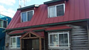 A two-story wooden house with a red roof, a front porch, and a window with a white frame.