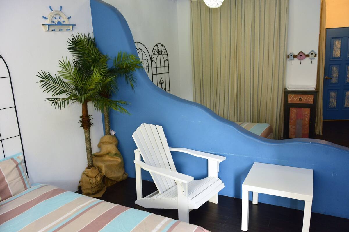 A room with a bed, a white chair, a small table, a palm tree, and a blue accent wall.