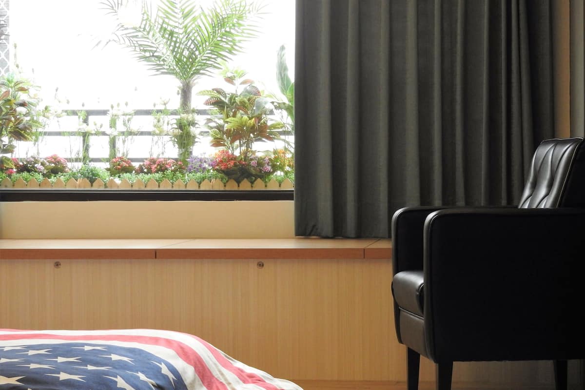 A room with a bed featuring an American flag design, a black leather chair, a window with a view of plants and a fence, and a wooden bench.
