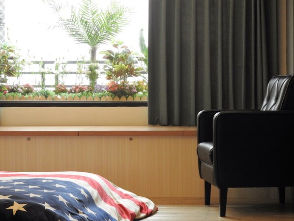 A room with a bed featuring an American flag design, a black leather chair, a window with a view of plants and a fence, and a wooden bench. - Anping House (Tainan)