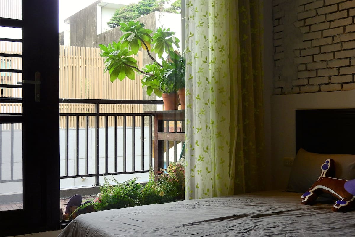 A bedroom with a bed, a window with a view of the street, a potted plant, and a curtain.