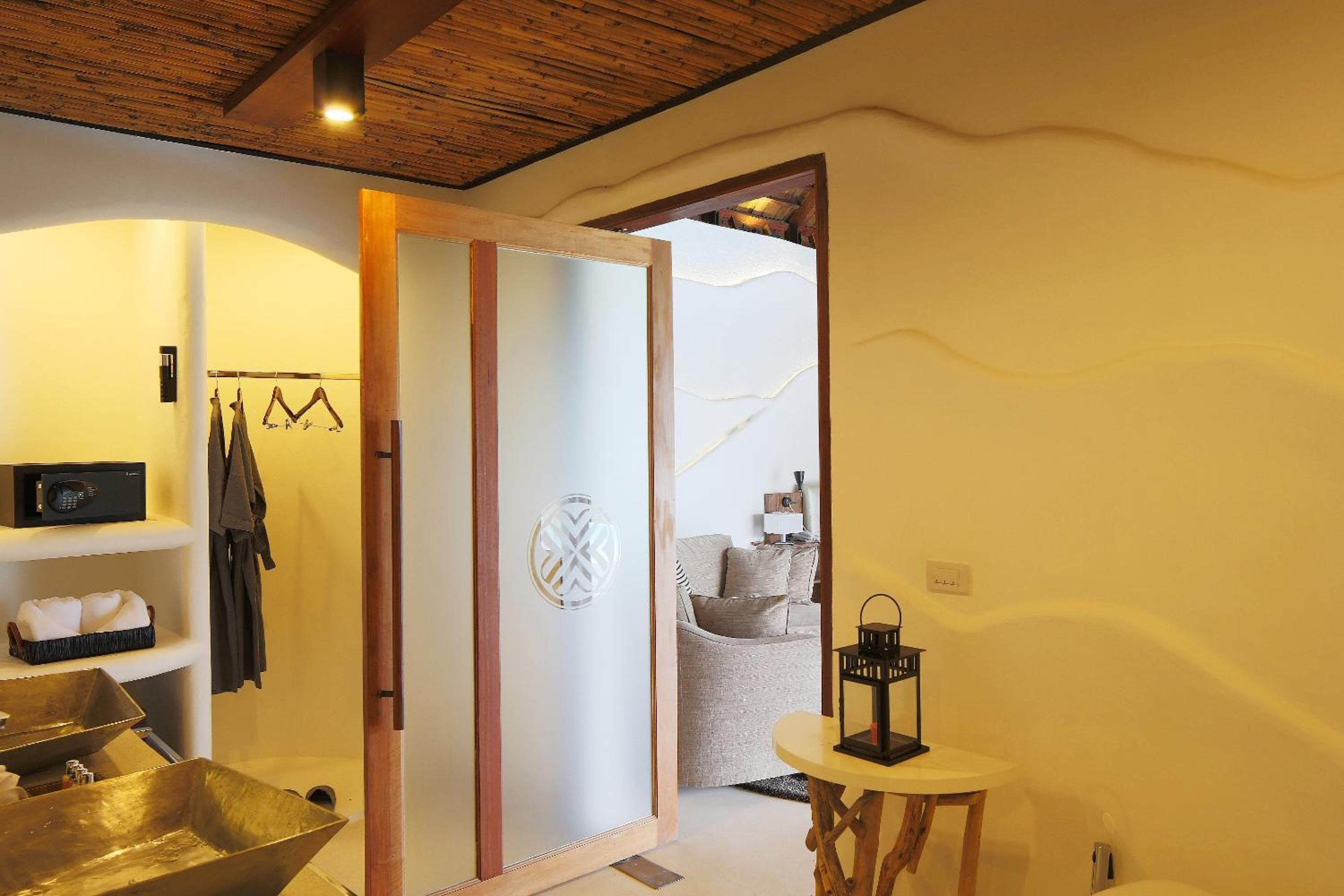 Una habitación con techo de madera, un espejo grande, un lavabo con pileta dorada, una mesa redonda pequeña con un farol, y una puerta que da a otra habitación.