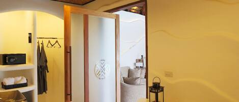 Una habitación con techo de madera, un espejo grande, un lavabo con pileta dorada, una mesa redonda pequeña con un farol, y una puerta que da a otra habitación.