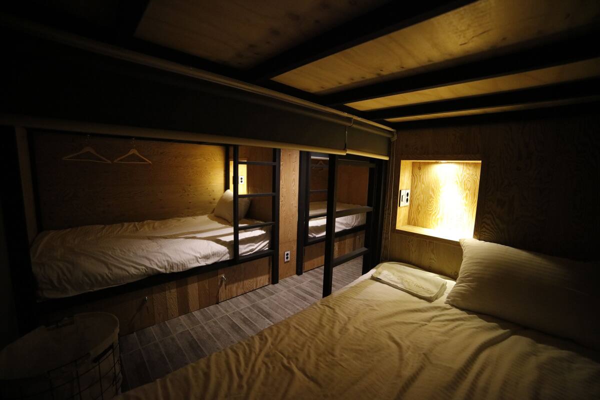 A dormitory-style room with bunk beds, a small table, and a window with a light fixture.