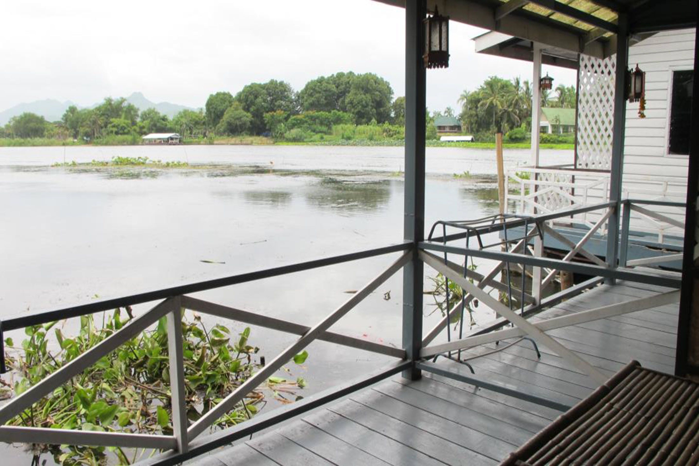 A covered outdoor area with a wooden deck, metal railing, and a view of a body of water with vegetation and distant buildings.
