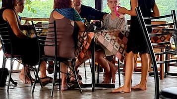 Un grupo de personas sentadas en una mesa al aire libre con piso de madera, rodeadas de sillas y una mesa cubierta con un mantel estampado.