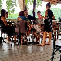 A group of people seated at an outdoor table with a wooden floor, surrounded by chairs and a table covered with a patterned tablecloth.