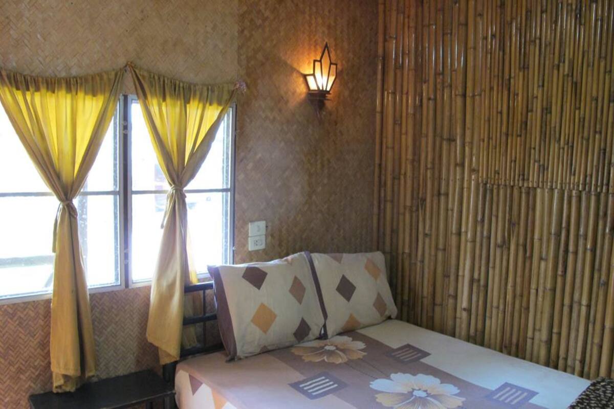 A room with a bed, two windows with yellow curtains, a wall-mounted light fixture, and a bamboo wall panel.