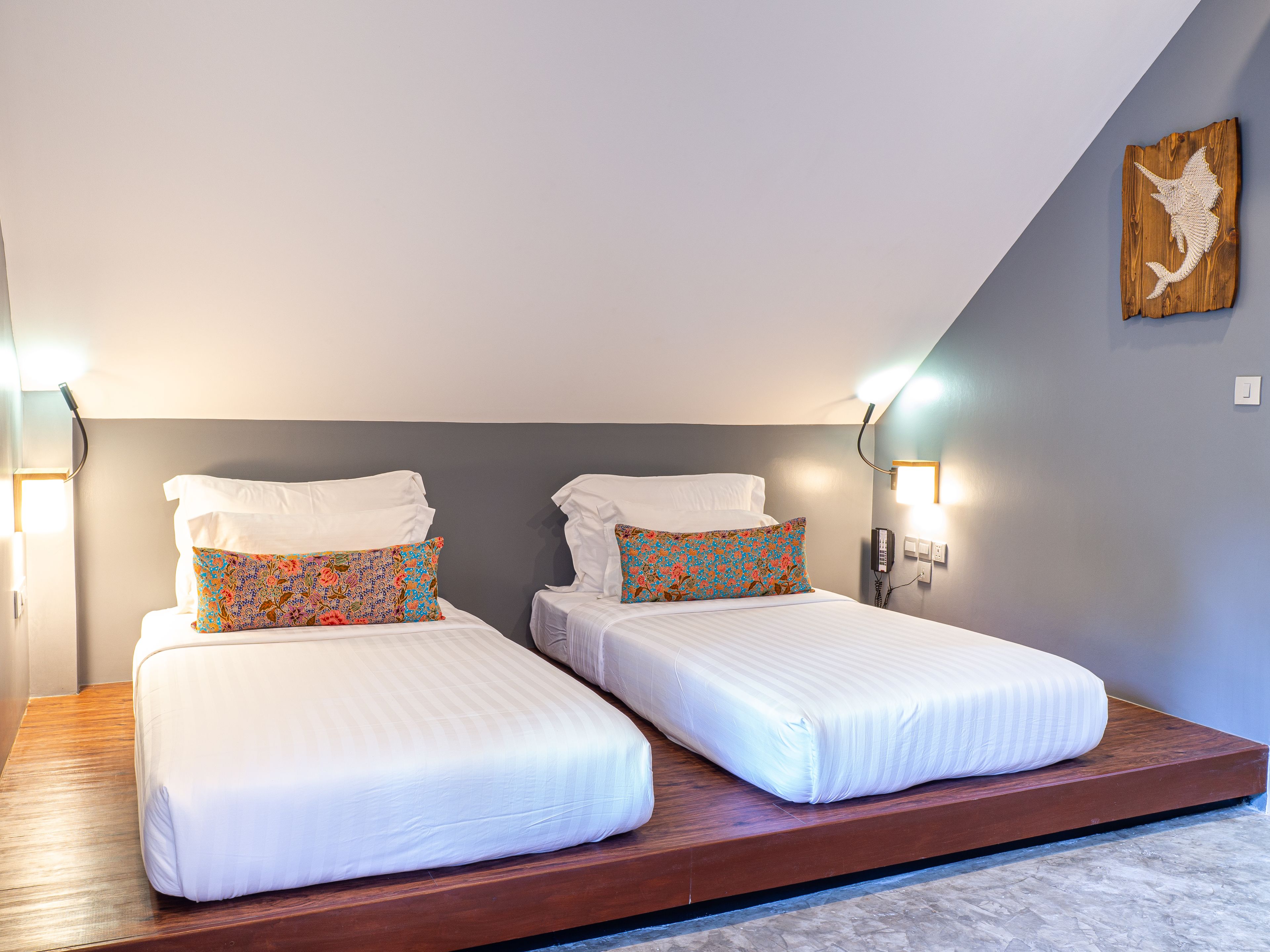 Une chambre dotée de deux lits simples, chacun agrémenté de draps blancs et d’oreillers à motifs, ainsi que d’une plateforme en bois et d’un luminaire fixé au mur.