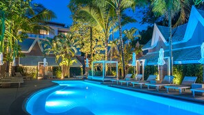 Ein Swimmingpool mit Liegestühlen und Sonnenschirmen, umgeben von Palmen und Gebäuden im traditionellen Stil.