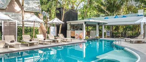 A swimming pool with lounge chairs and umbrellas, surrounded by trees and a covered area with a bar.