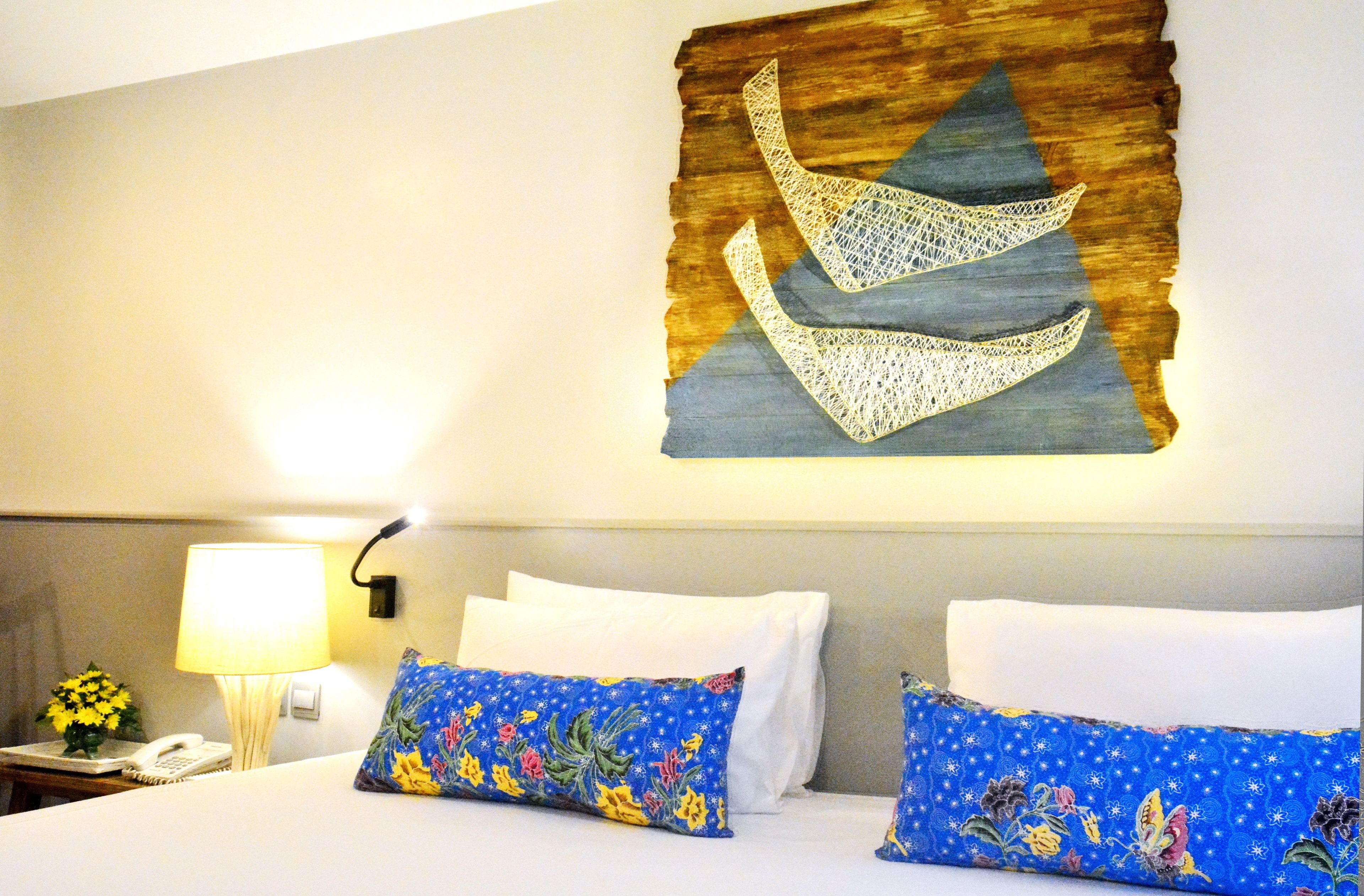 Habitación de hotel con cama, almohadas blancas, una almohada con estampado floral azul, lámpara, teléfono y una obra de arte en la pared.