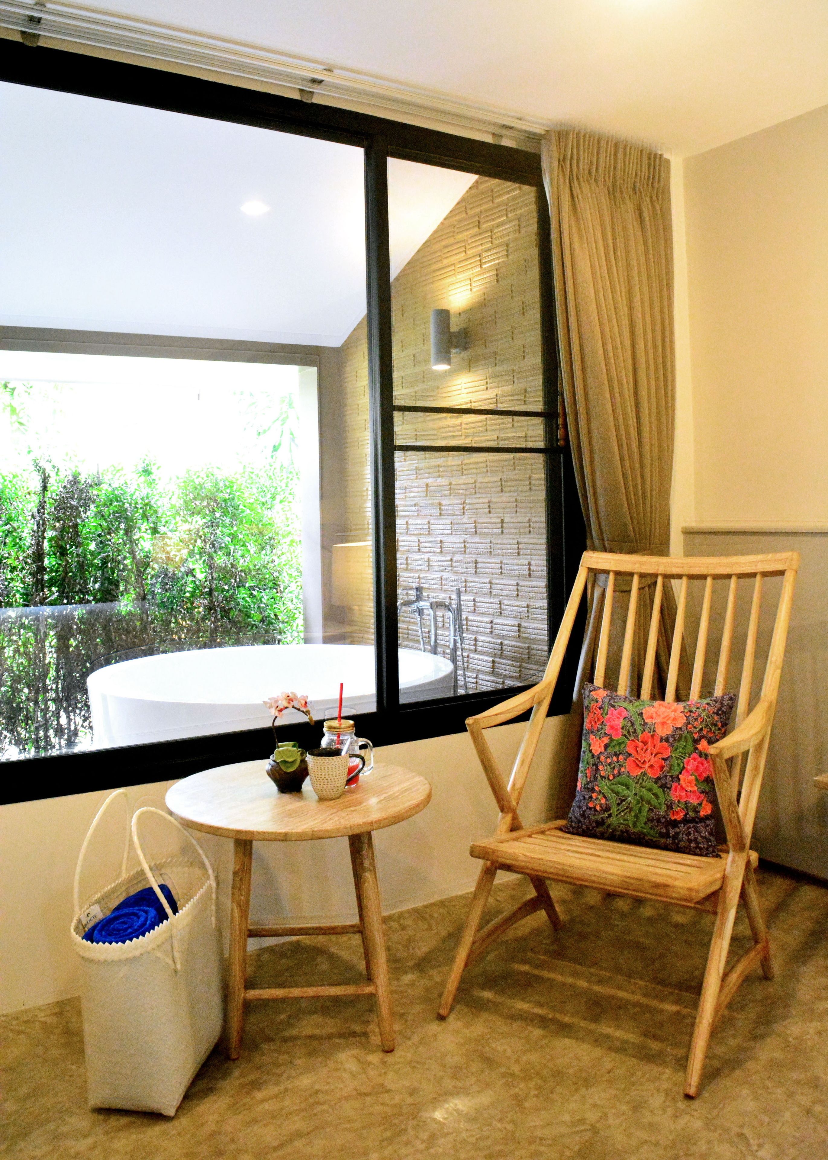 A wooden chair with a floral pillow, a small round table with a drink and a flower, and a white bag on the floor.