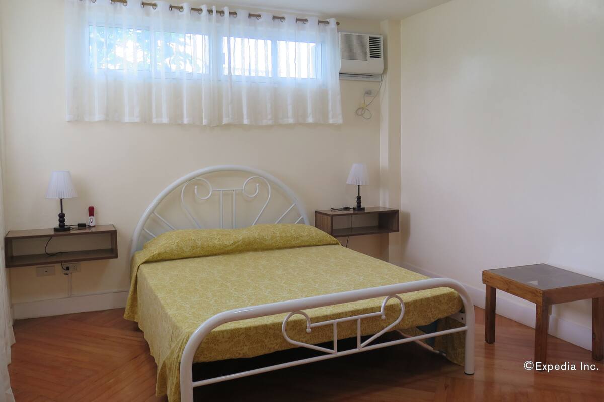 Un lit simple avec un cadre en métal et une couvre-lit jaune, deux tables de chevet avec des lampes, une petite table d’appoint en bois et un climatiseur fixé au mur.