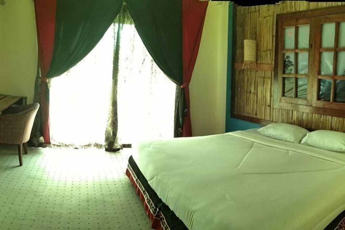 A room with a bed, a window with curtains, a chair, and a wall-mounted lamp.