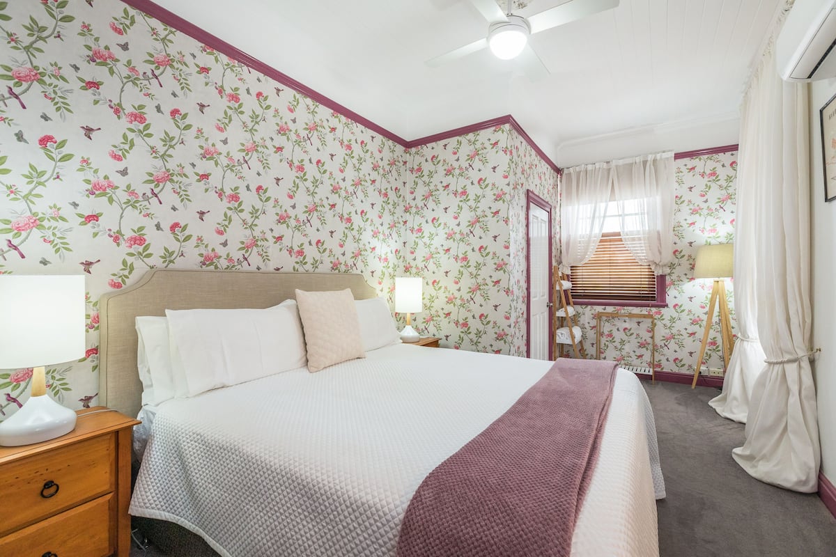 Ein Schlafzimmer mit blumigem Wandpapier, einem Bett mit weißer und pinkfarbener Bettwäsche, einem Nachttisch mit Lampe und einer Holzleiter.