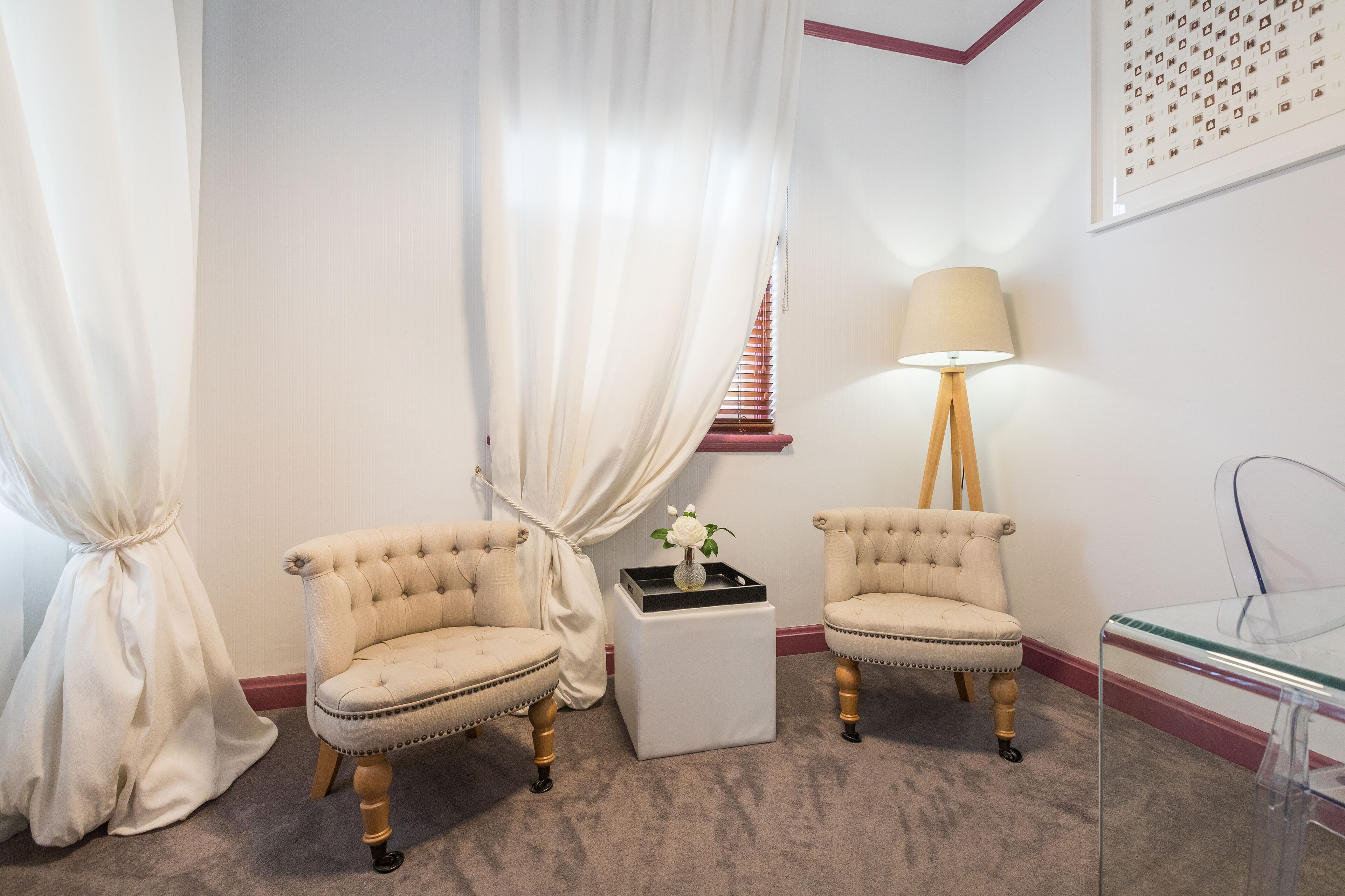 A room with two upholstered armchairs, a small white side table with a vase of flowers, a floor lamp, and a glass table.