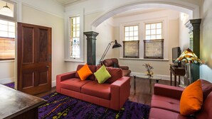 Un salón con un sofá rojo, una mesa de centro y una lámpara. Hay ventanas con persianas, una puerta y una escalera que lleva a un nivel superior.