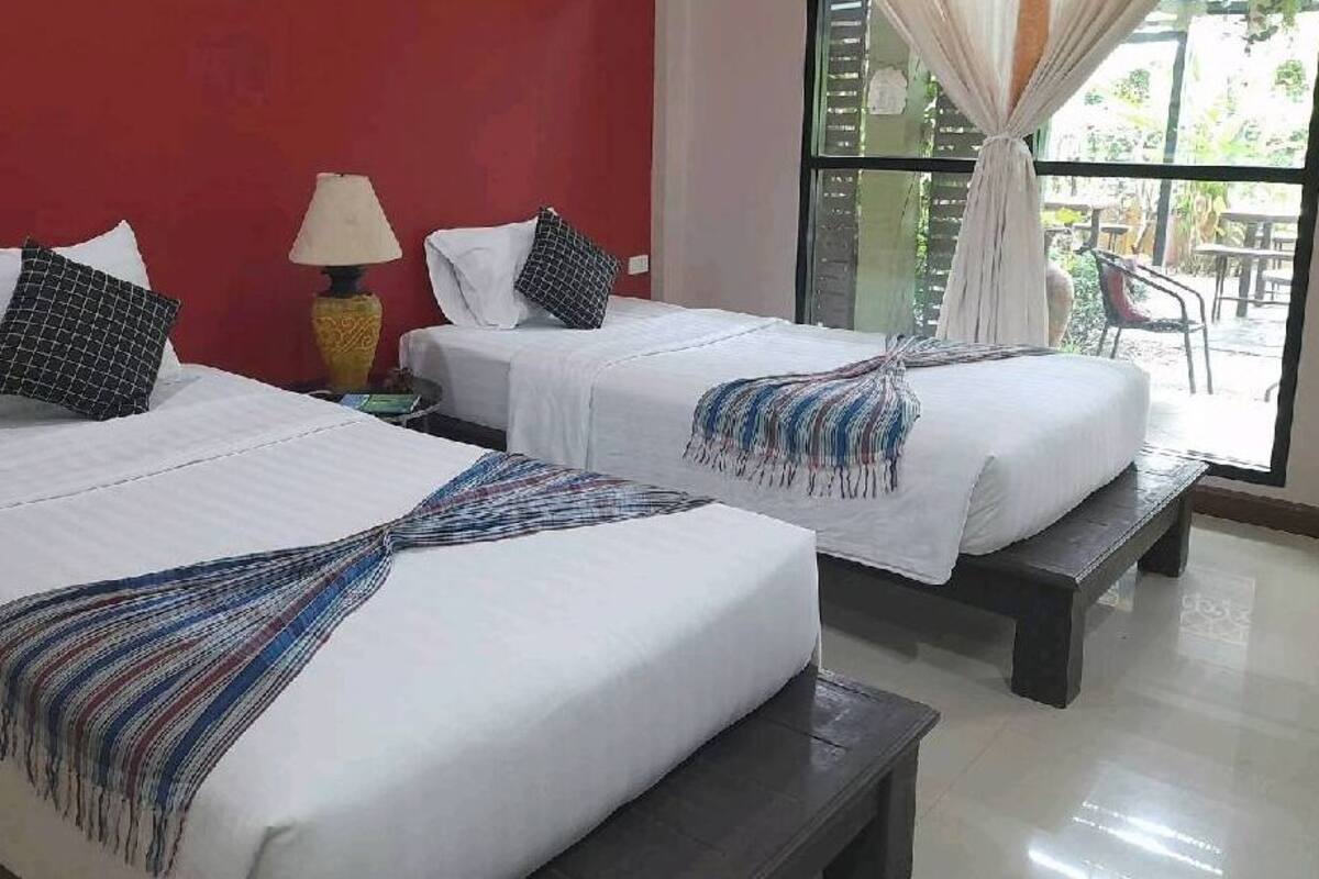 Dos camas individuales con ropa de cama blanca y almohadas estampadas, una mesita de noche con lámpara, y vista al exterior a través de una ventana con cortinas.