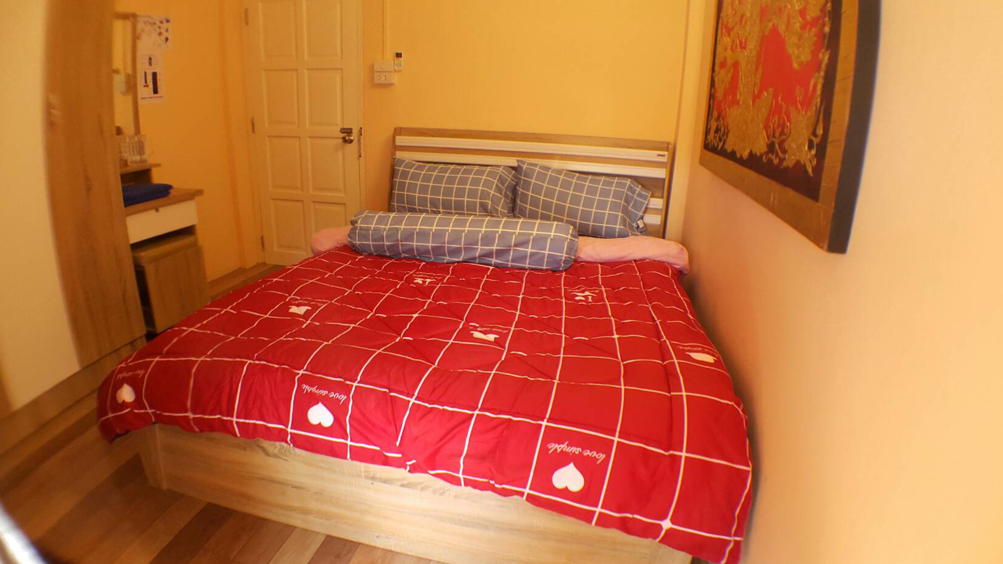 Quarto com estrutura de cama de madeira, colcha xadrez vermelha e branca, e travesseiro azul e branco.
