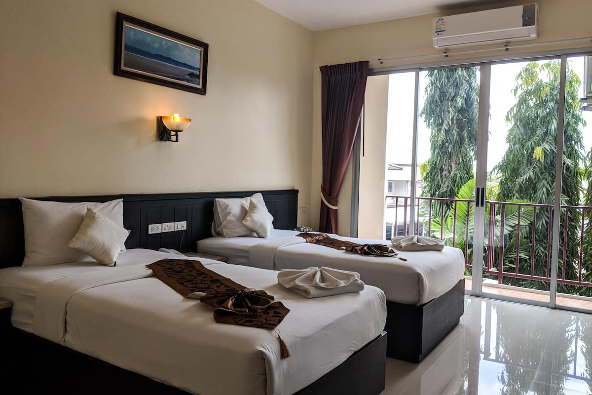 A hotel room with two beds, a large window with a view of greenery, an air conditioning unit, and a framed picture on the wall.