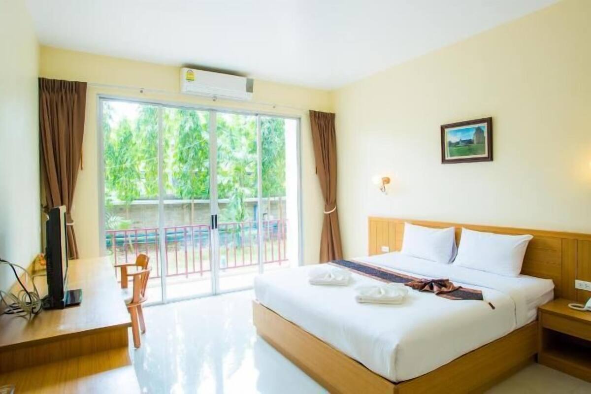 A hotel room with a large bed, a desk with a chair, a bedside table with a phone, and a view of greenery outside.